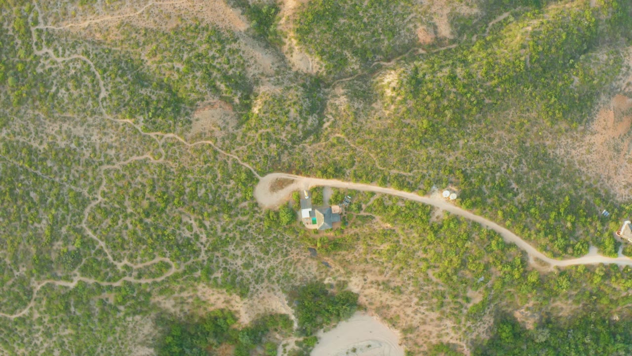 Cabo Pulmo overhead frame showing the winding dirt road across scrubby hills, a small light vehicle on the roadway, nearby structures, and no visible people.