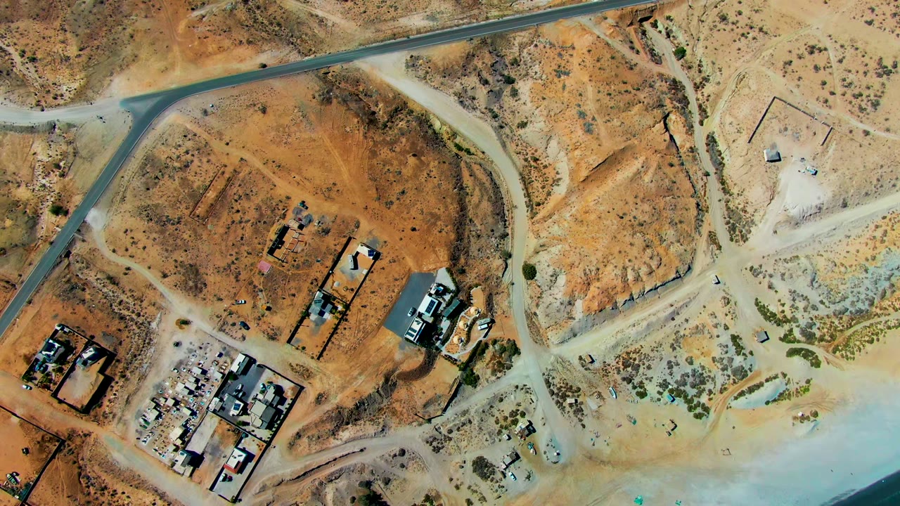 Overhead drone frame in San Juanico, Baja California Sur showing dirt roads, scattered structures, cleared desert ground, beach sand, pale water, and no visible people.
