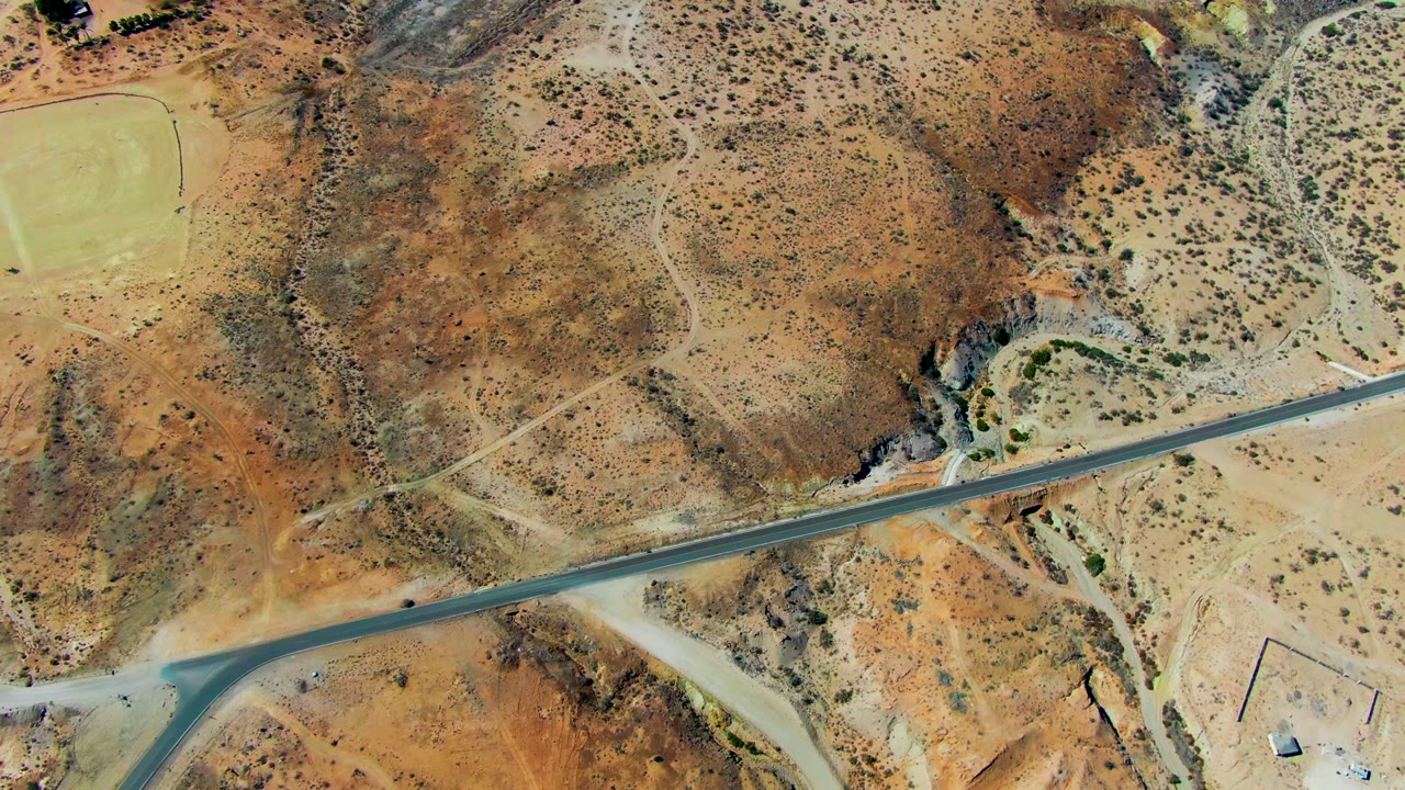 The San Juanico aerial view keeps the runway running across the lower frame, bordered by tan dirt, rust hills, scattered brush, and no visible people.