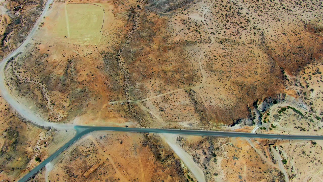 Overhead view in San Juanico, Baja California Sur of a paved road meeting pale dirt tracks across rust and tan desert hills, with sparse scrub and no visible people.