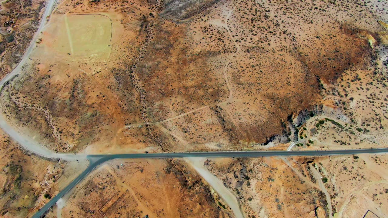 Overhead view of San Juanico, Baja California Sur, showing a paved road across dry terrain, a pale green field near the upper left, dirt paths, scrub, and no visible people.
