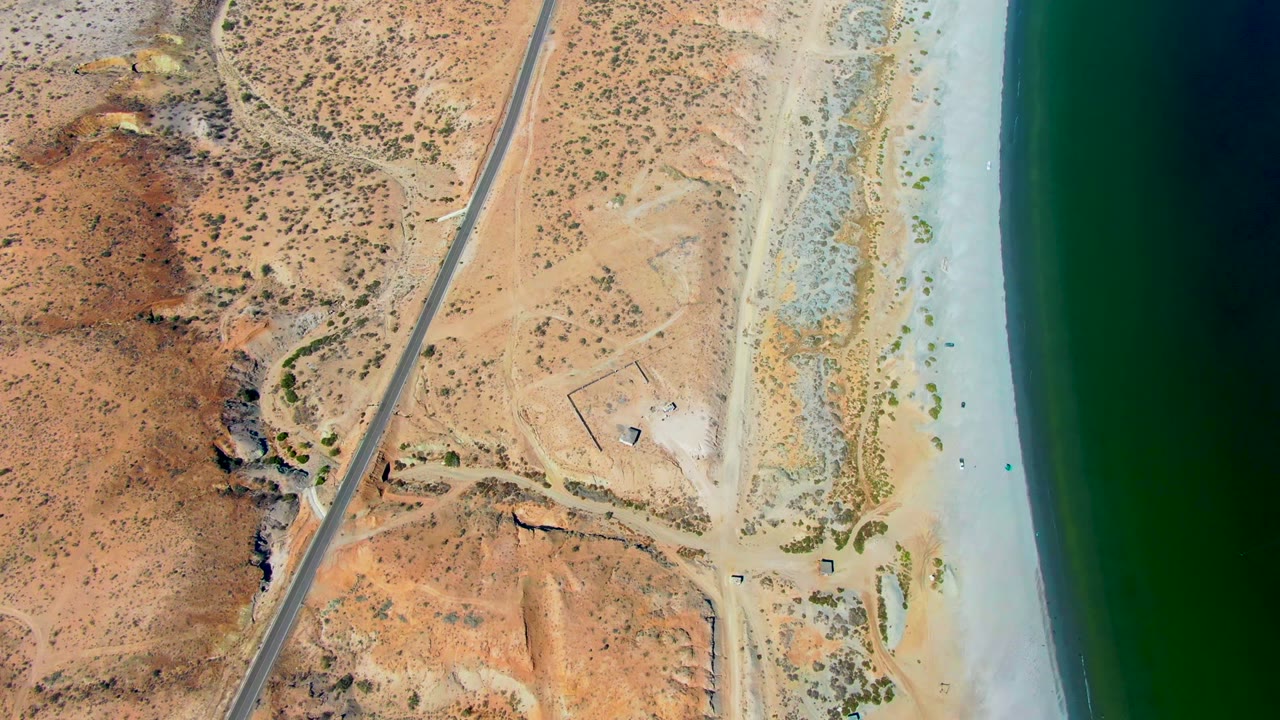 Overhead drone-style view in San Juanico, Baja California Sur, with a paved road cutting through dry scrubland beside pale beach and green ocean water, no visible people.