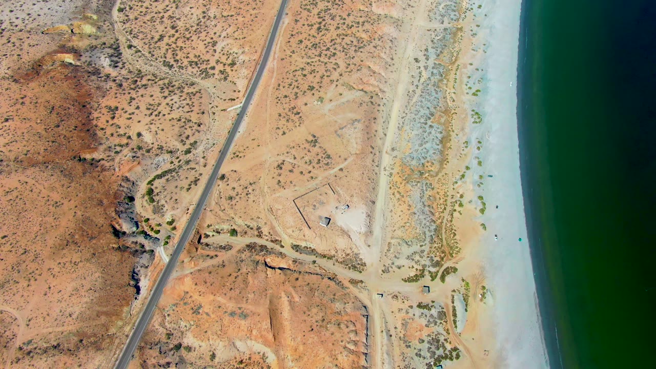 Aerial coastal road crossing arid terrain near a sandy shoreline.