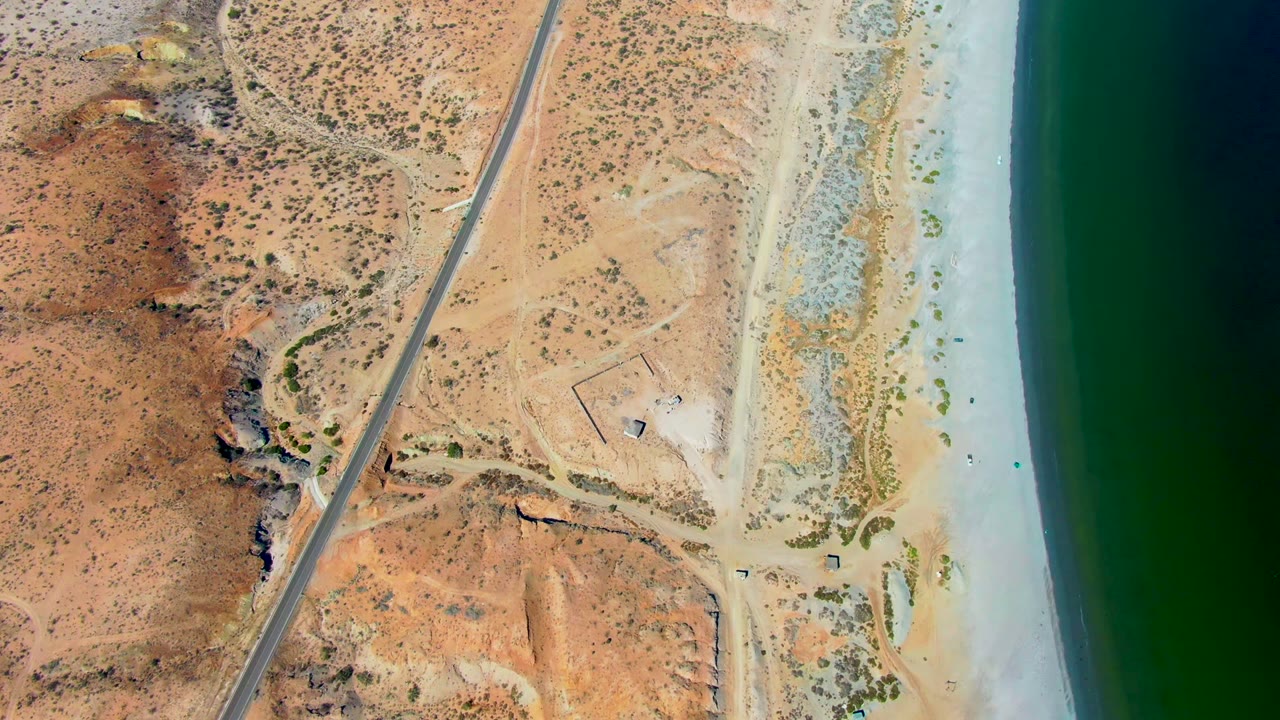 High overhead frame in San Juanico showing reddish desert hills on the left, a straight road near the shore, pale beach flats, and green ocean water on the right, no visible people.