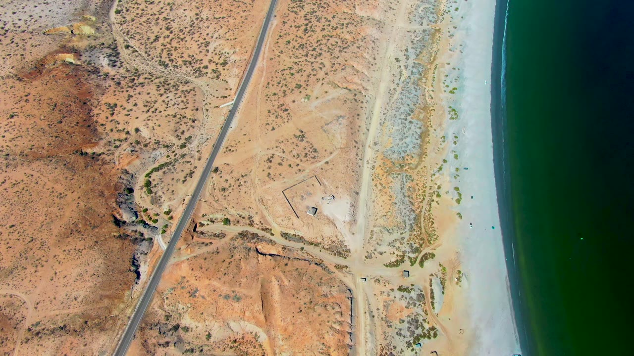 High aerial frame over San Juanico, Baja California Sur showing tan desert scrub, a narrow road, pale beach, dark green ocean, and no visible people.