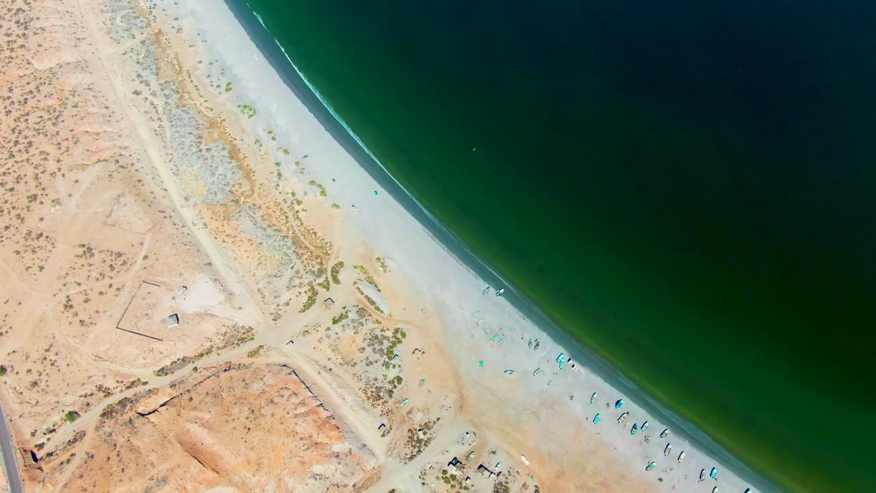 Top-down San Juanico coast frame with dark green ocean filling the right side, pale beach below the desert edge, scattered umbrellas or small beach setups, and tiny people near the waterline.