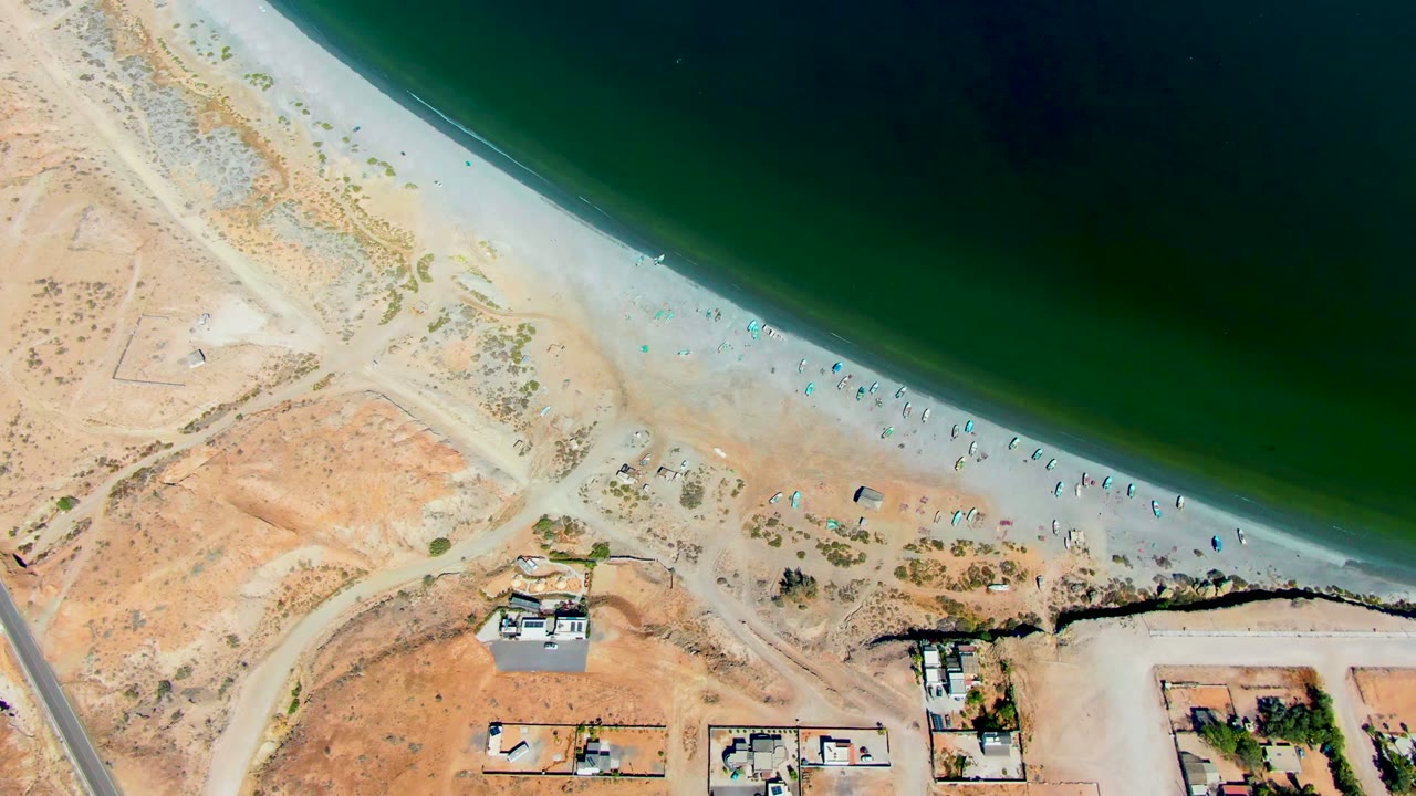 Aerial frame over San Juanico, Baja California Sur, with dark green water at the top, pale beach through the middle, scattered tiny beachgoers, and coastal buildings near the lower edge.