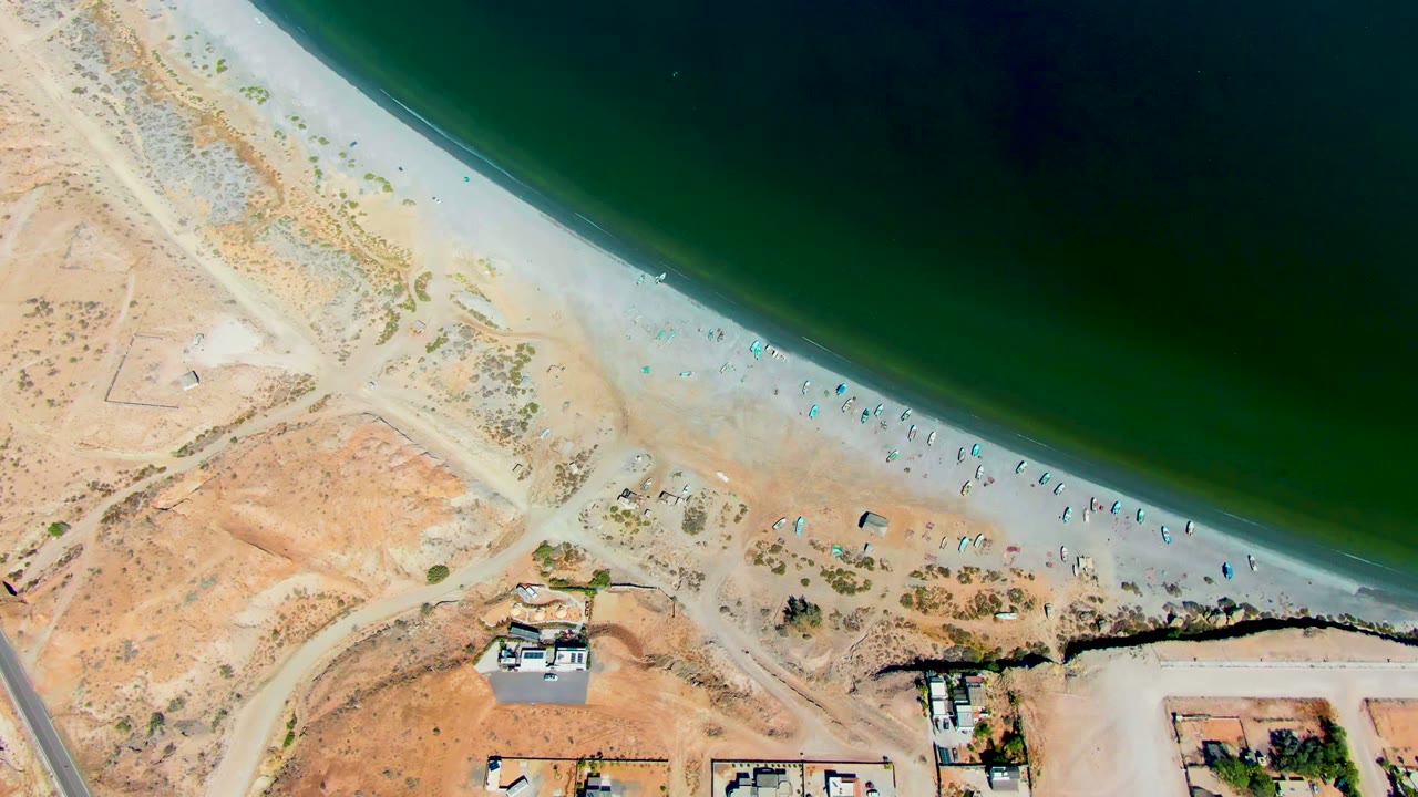Overhead coastline with pale sand, green water, beachgoers, desert tracks, road, and small buildings.