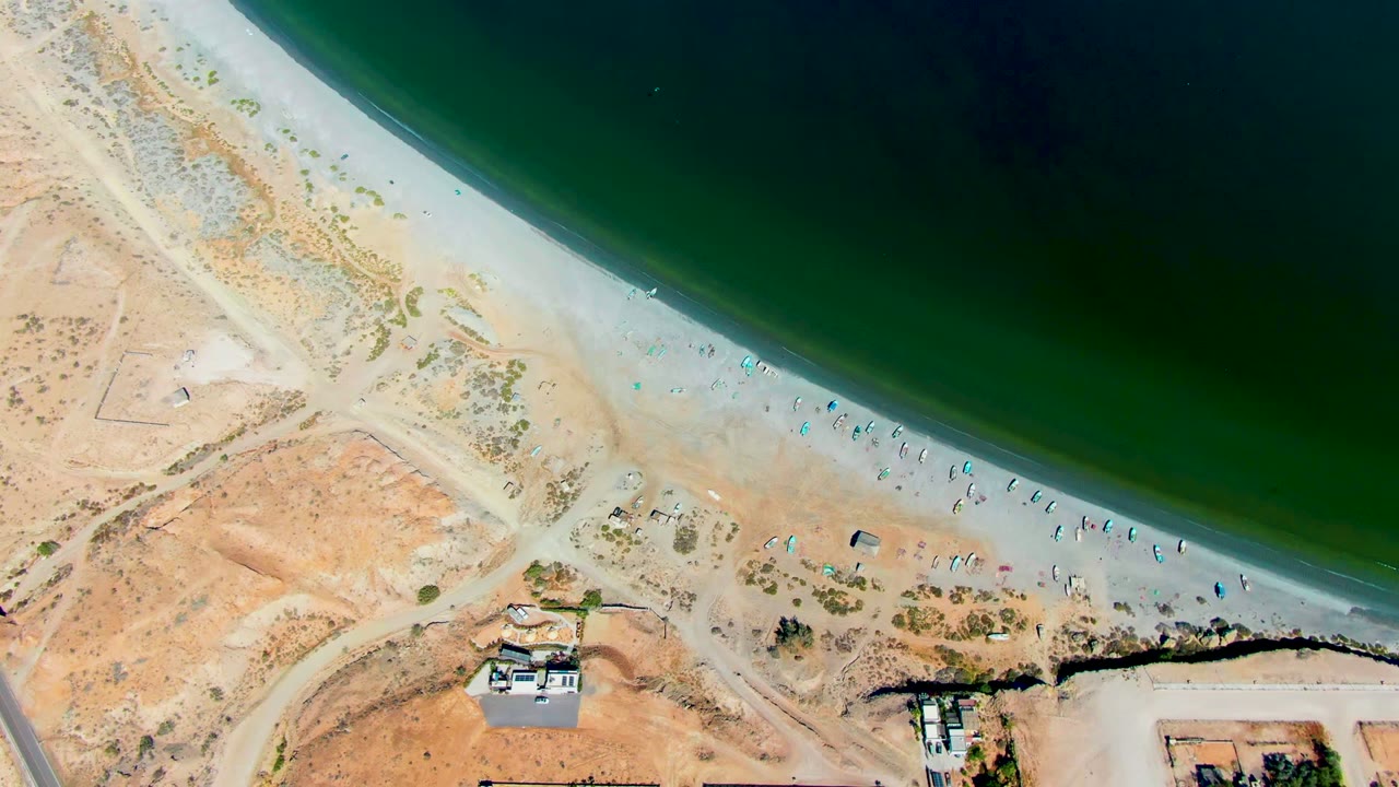 San Juanico aerial frame with the ocean occupying the upper right, a narrow bright beach lined with tiny visitors, and dry inland terrain crossed by a road and dirt access tracks.