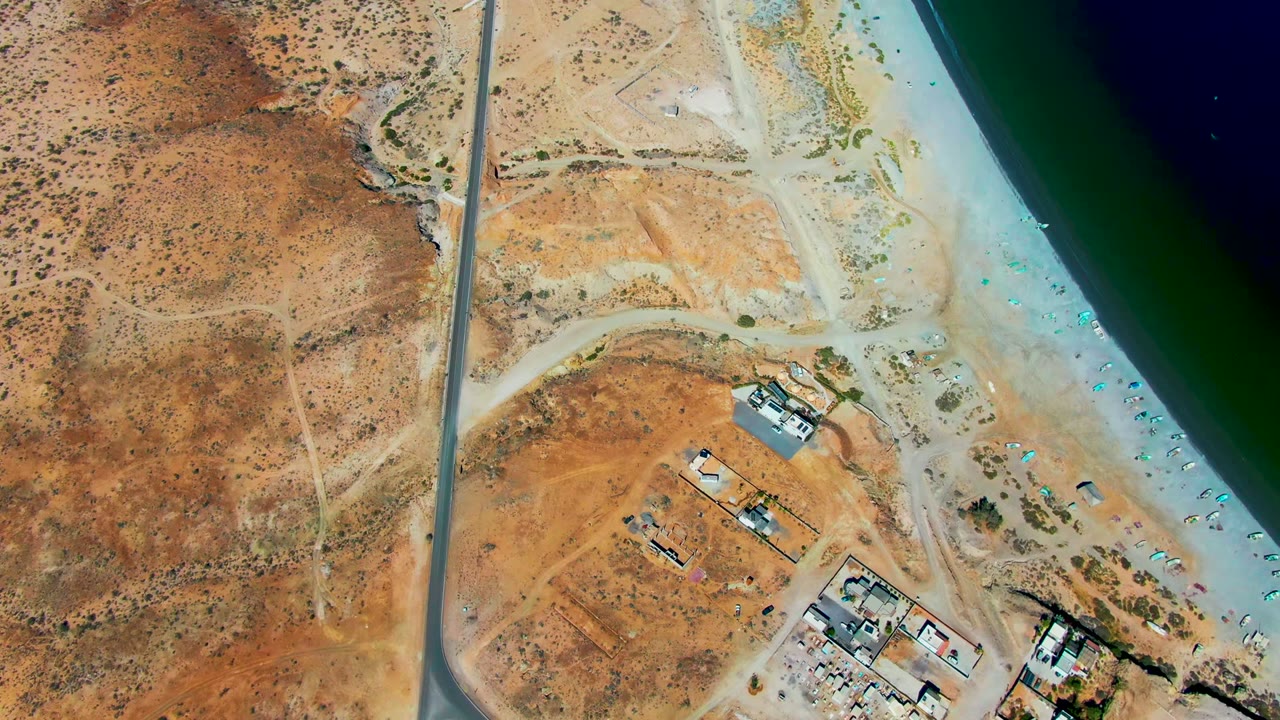 Aerial frame over San Juanico, Baja California Sur showing a paved road beside arid land, sandy beach, green water, and small coastal structures, with no visible people.