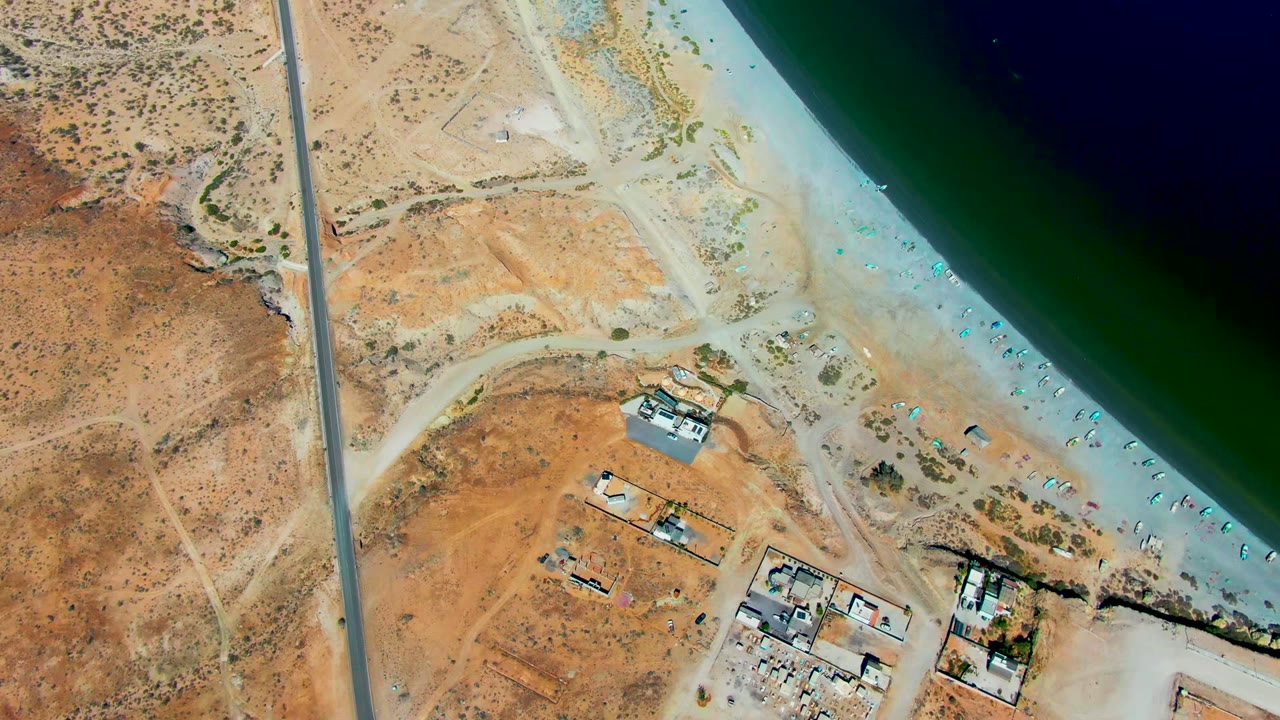 High aerial view of dry coastal land, road, beach, water, and small buildings.