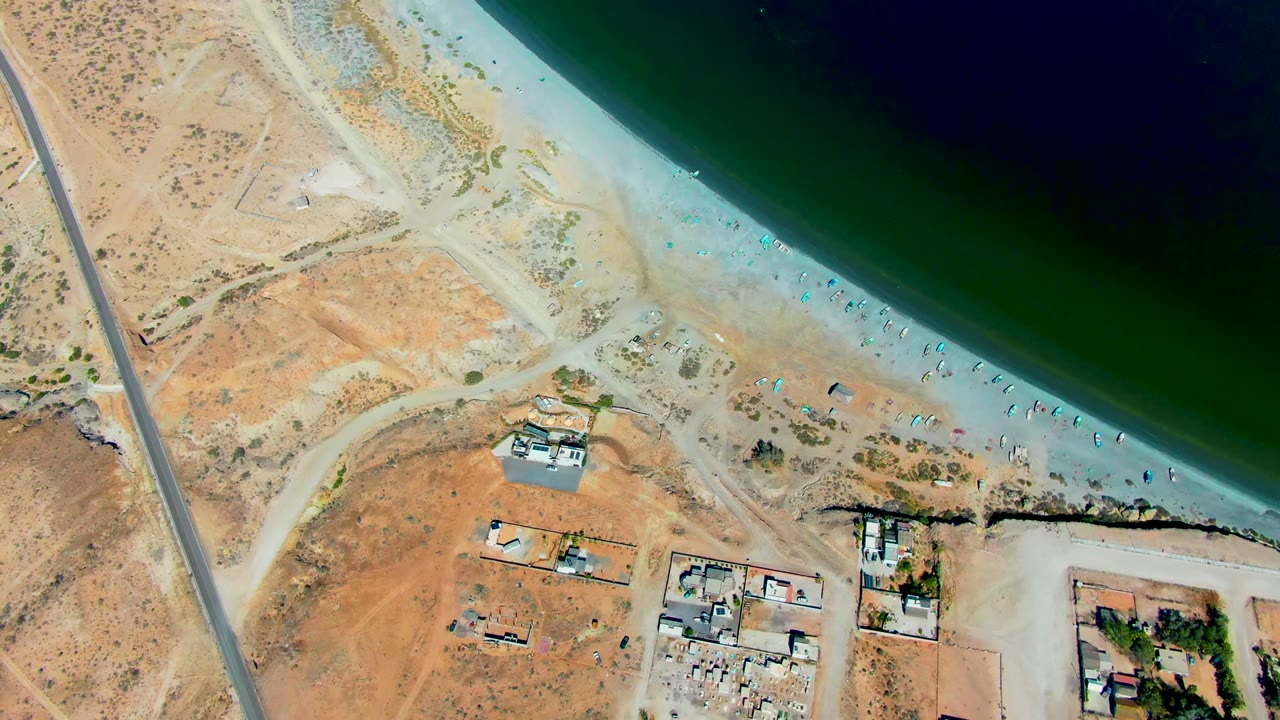 Aerial frame of San Juanico coastline where the curving beach and green water dominate the top of frame, with dry land, access tracks, and small buildings below, with no visible people.