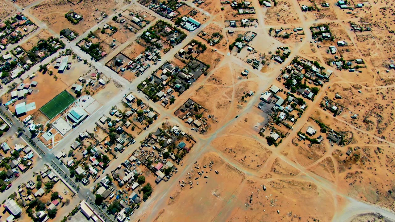 Overhead view of San Juanico residential blocks, pale roads, open dirt lots, and scattered houses in arid terrain, with no visible people.