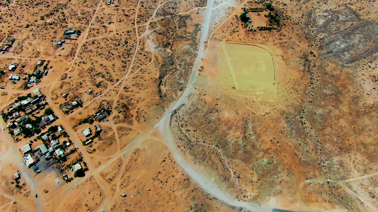 High overhead view in San Juanico with residential clusters, winding dirt tracks, and broad tan open land, with no visible people.