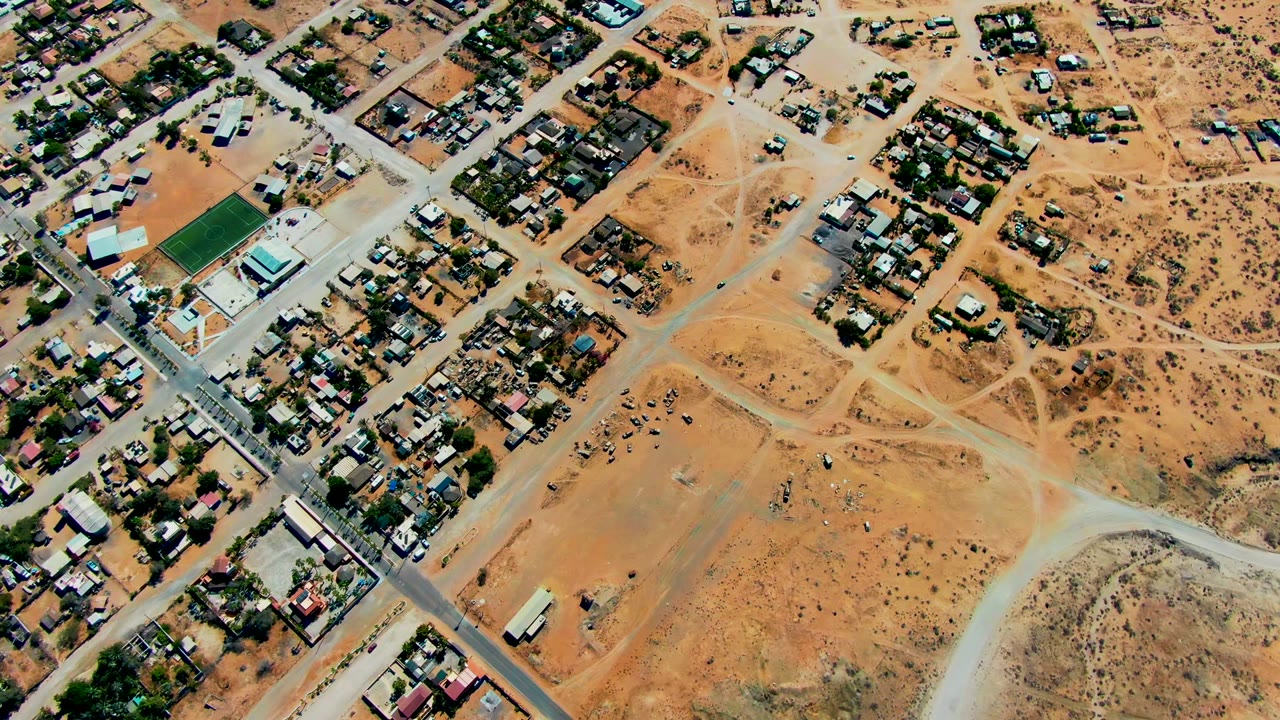 Overhead frame of San Juanico, Baja California Sur, with tan desert lots, clustered homes, paved roads, dirt tracks, a green sports field at left, and no visible people.