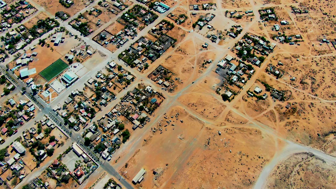 High overhead frame of San Juanico with residential blocks, a diagonal paved road, pale dirt paths through dry ground, scattered vehicles, and no visible people.