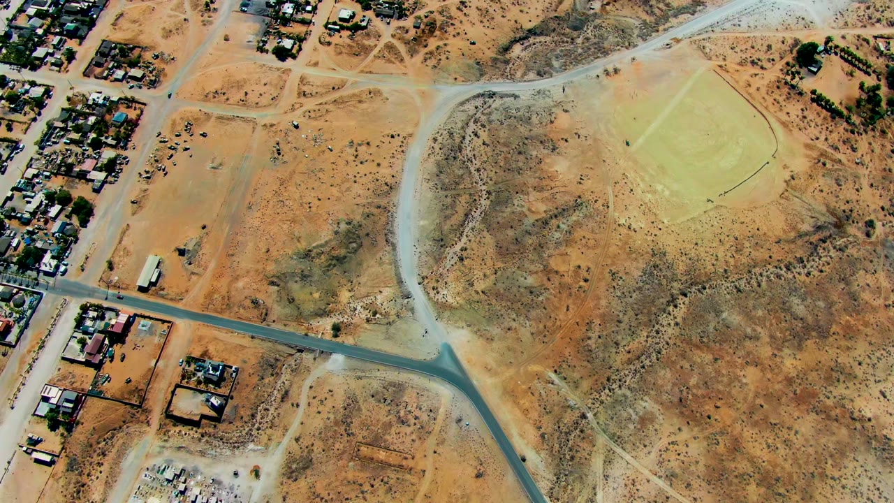 Overhead drone frame of San Juanico, Baja California Sur, showing tan desert ground, scattered homes, dirt tracks, a pale sports field, and no visible people.