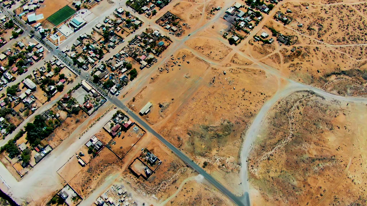 Aerial view in San Juanico with a cluster of homes at the upper left, a diagonal paved road, branching dirt tracks, orange desert lots, and no visible people.