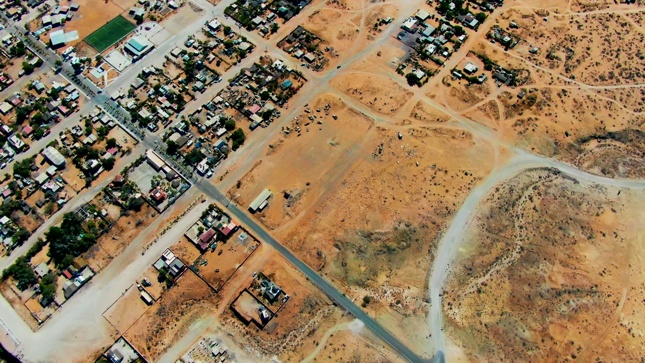 San Juanico aerial frame showing houses along the top edge, a gray road crossing the scene, pale dirt access roads, dry lots, and no visible people.