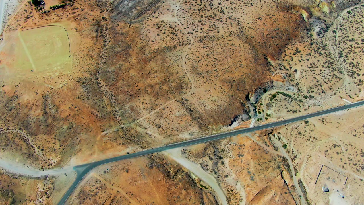Aerial view over San Juanico, Baja California Sur, showing a paved road, pale dirt junctions, brown scrubland, rocky washes, and a small green field, with no visible people.