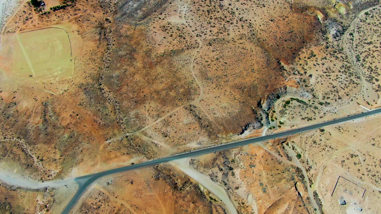Aerial San Juanico frame with a straight paved road below, brown desert hills across the center, a rocky drainage wash to the right, and a pale green field near the upper left, with no visible people.