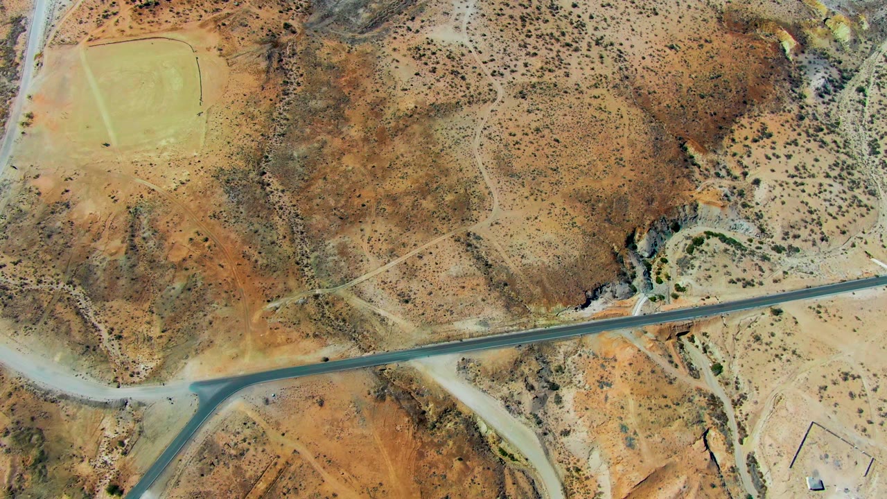 High aerial view near San Juanico, Baja California Sur, with a paved road crossing dry ocher terrain, a T-junction at lower left, sparse scrub, and no visible people.