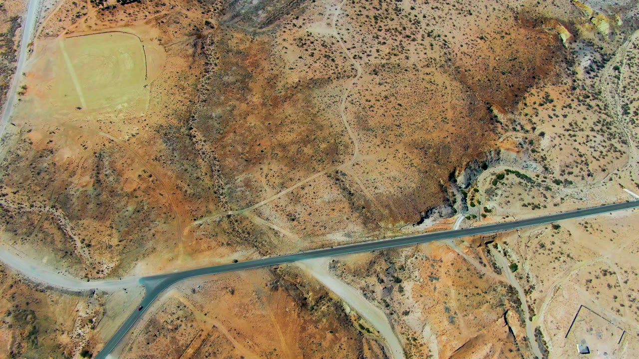 Overhead aerial frame in San Juanico, Baja California Sur, showing a paved road across dry brown terrain, pale dirt tracks, sparse scrub, and no visible people.