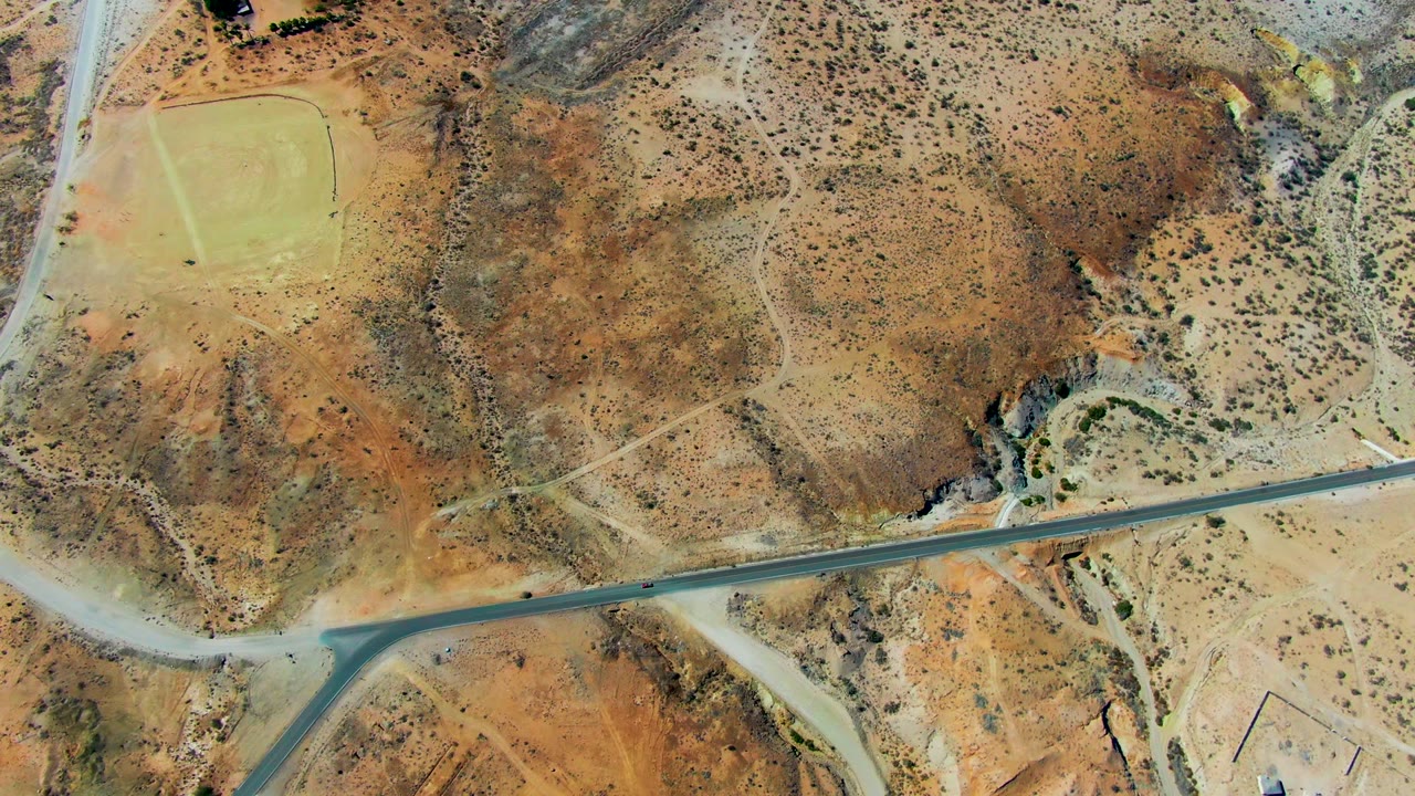 High aerial view near San Juanico, Baja California Sur, showing a small vehicle entering a straight desert road area bordered by reddish earth, scrub, dirt tracks, and no visible people.