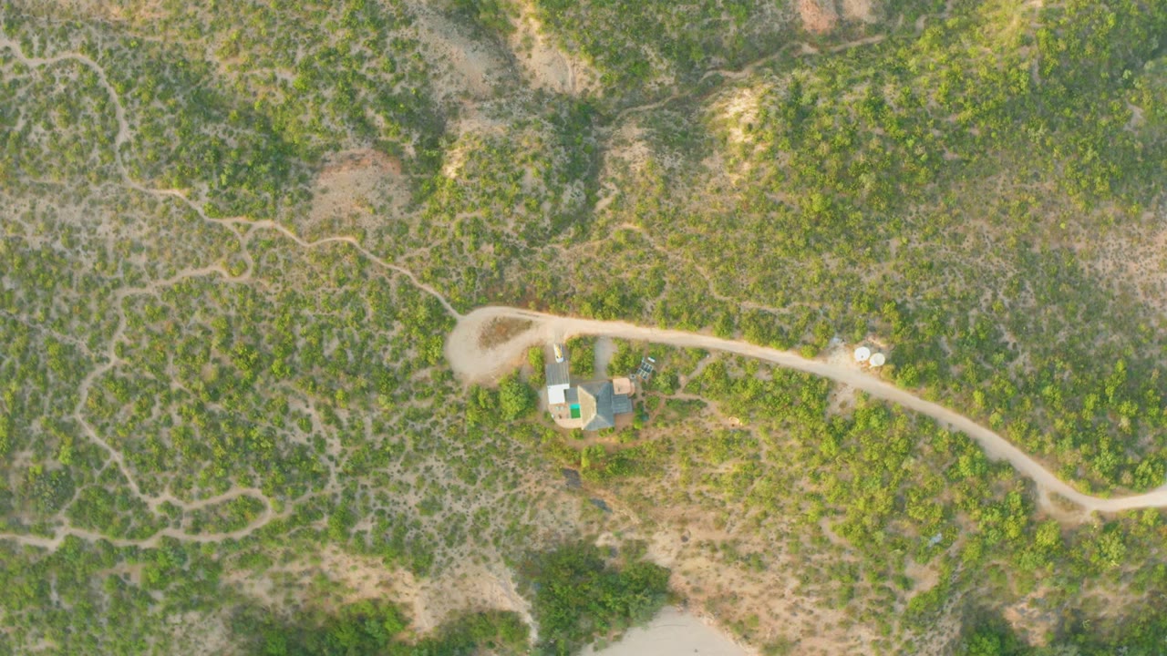 Overhead aerial frame in Cabo Pulmo, Baja California Sur, showing a pale dirt road curving past small roofed buildings amid green and brown scrub, with no visible people.