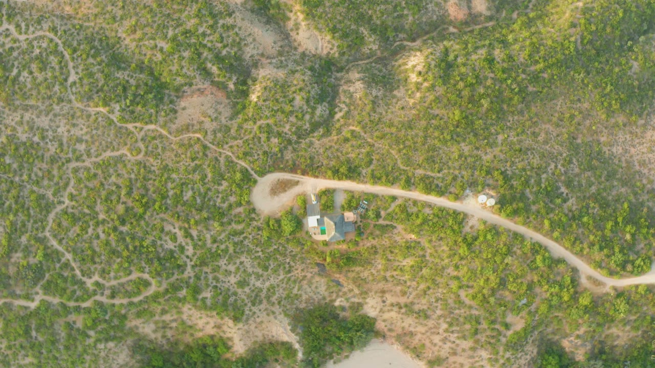 Aerial view of a winding dirt road beside small structures in scrubland.