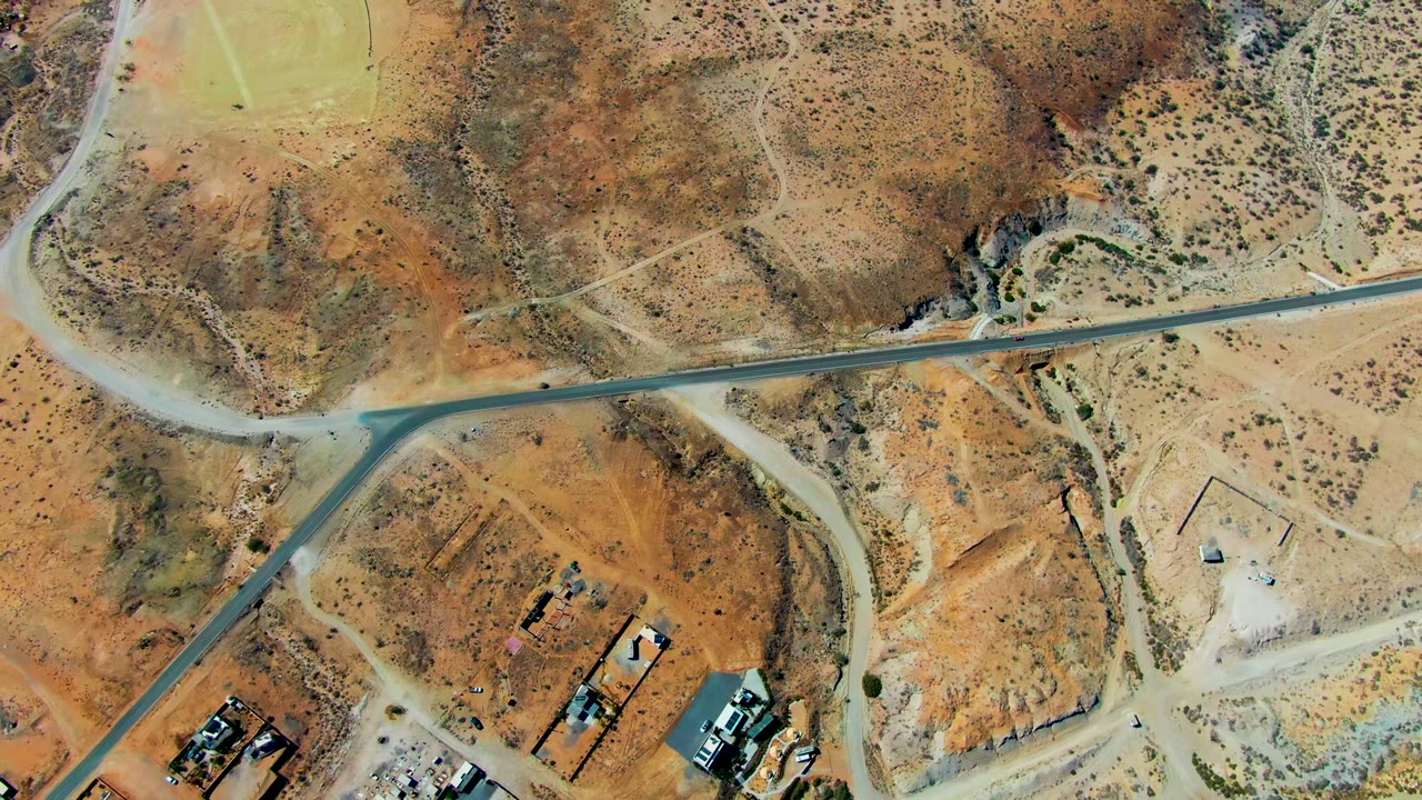 Overhead view of San Juanico desert terrain with a paved road, pale dirt tracks, scrub vegetation, small structures at the lower edge, and no visible people.