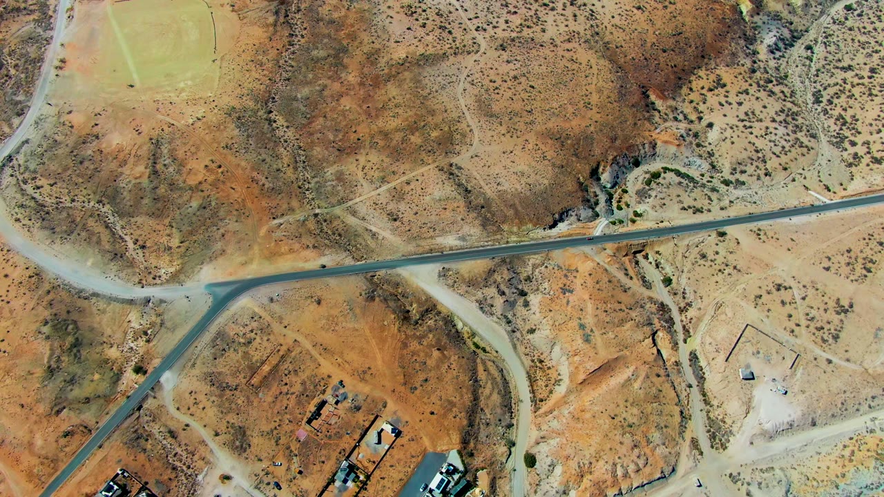 San Juanico drone view with the paved road across the middle, dry washes and scrub above it, small structures and vehicles near the lower edge, and no visible people.