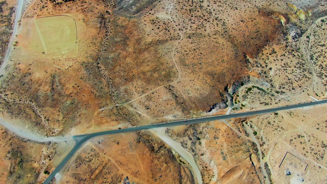 Overhead view in San Juanico, Baja California Sur showing a straight road crossing dry brown terrain, a small green field at upper left, scattered scrub, and no visible people.