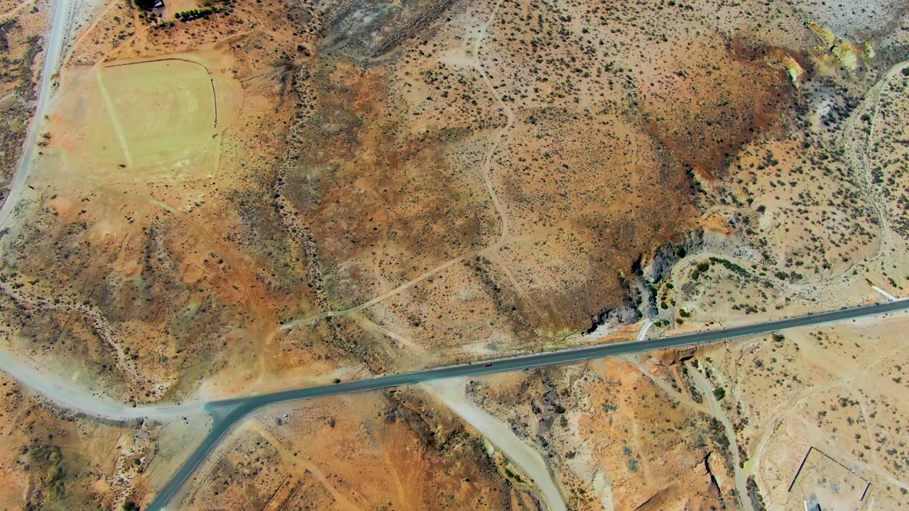 Top-down San Juanico, Baja California Sur view showing the road stretched across the bottom third, dry washes and scrub above it, the small vehicle on the road, and no visible people.