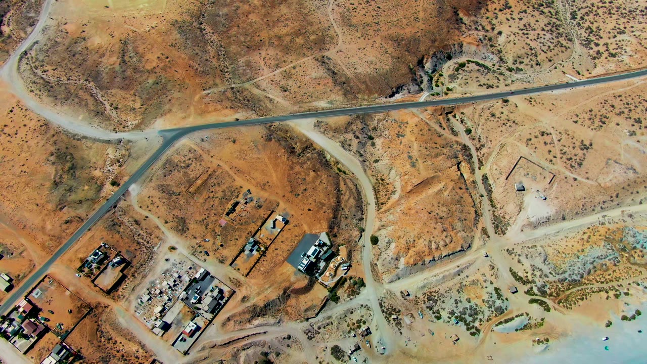 Overhead view in San Juanico, Baja California Sur, showing a paved road crossing dry brown terrain with dirt tracks, scrub, building clusters, and no visible people.