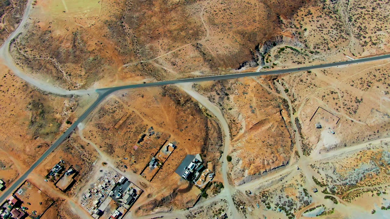Overhead San Juanico frame with a winding paved road, orange-brown hills, pale cleared rectangles, scattered structures, and no visible people.