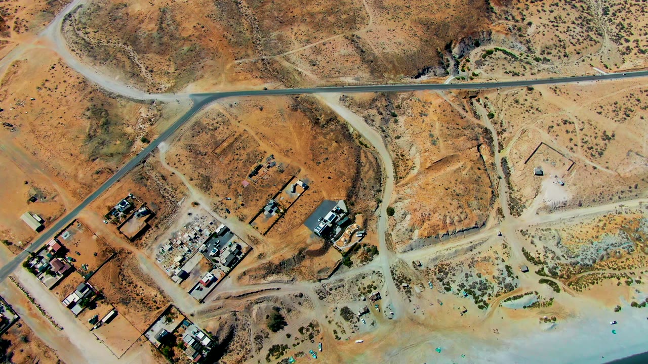 High aerial frame over San Juanico, Baja California Sur, with dirt roads, dry hills, clustered structures and vehicles at lower left, shoreline at the bottom edge, and no visible people.