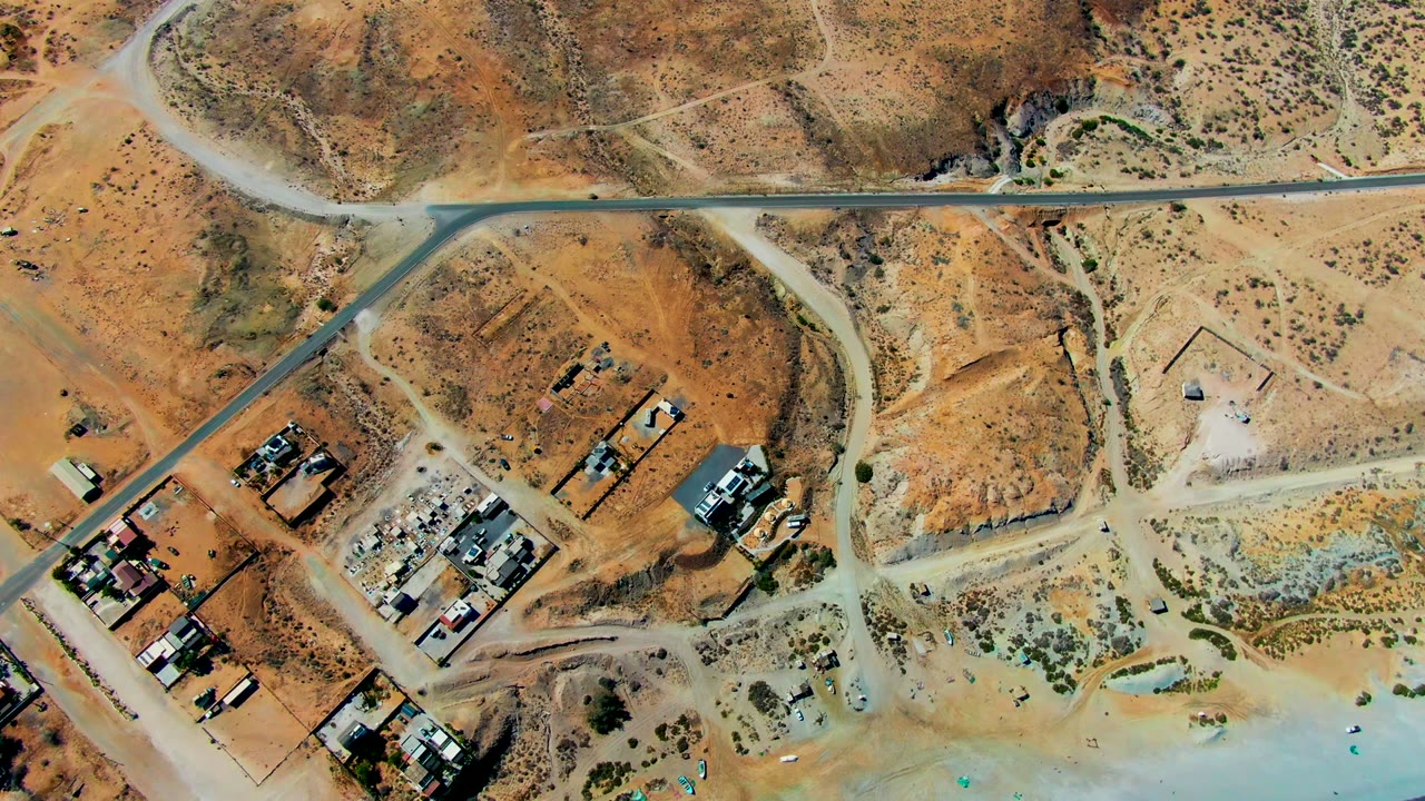Overhead view in San Juanico, Baja California Sur showing a paved road, branching dirt tracks, dry orange terrain, scattered structures, scrub, and no visible people at the opening frame.