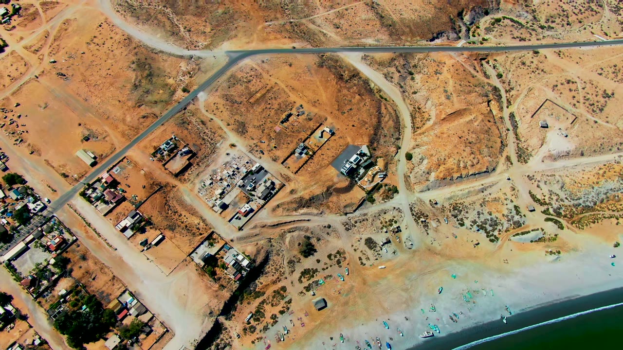 Overhead aerial frame of San Juanico, Baja California Sur showing dirt roads, scattered buildings, dry lots, pale beach, turquoise shoreline water, and no visible people.