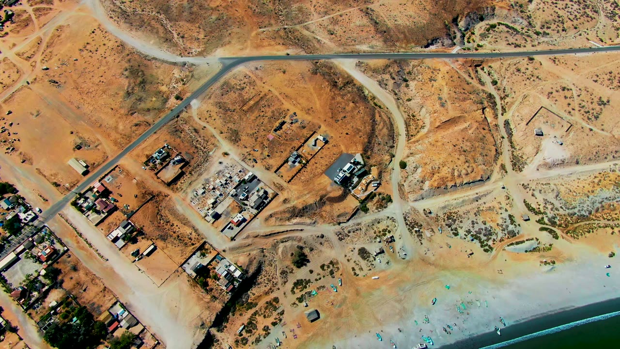 Drone view of dry coastal lots, a road crossing the frame, buildings, sand, umbrellas, and water.