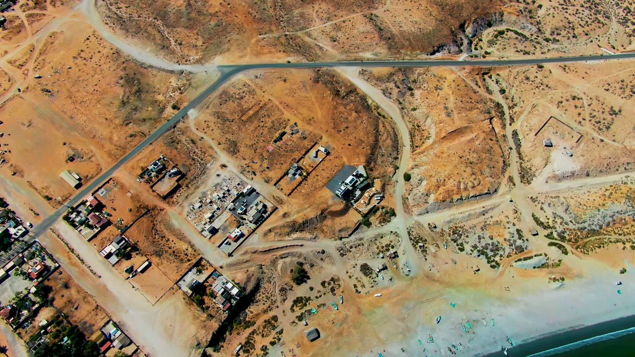 Top-down frame of San Juanico showing construction pads and dirt tracks above a sandy beach with blue umbrellas and a narrow band of water, with no visible people.