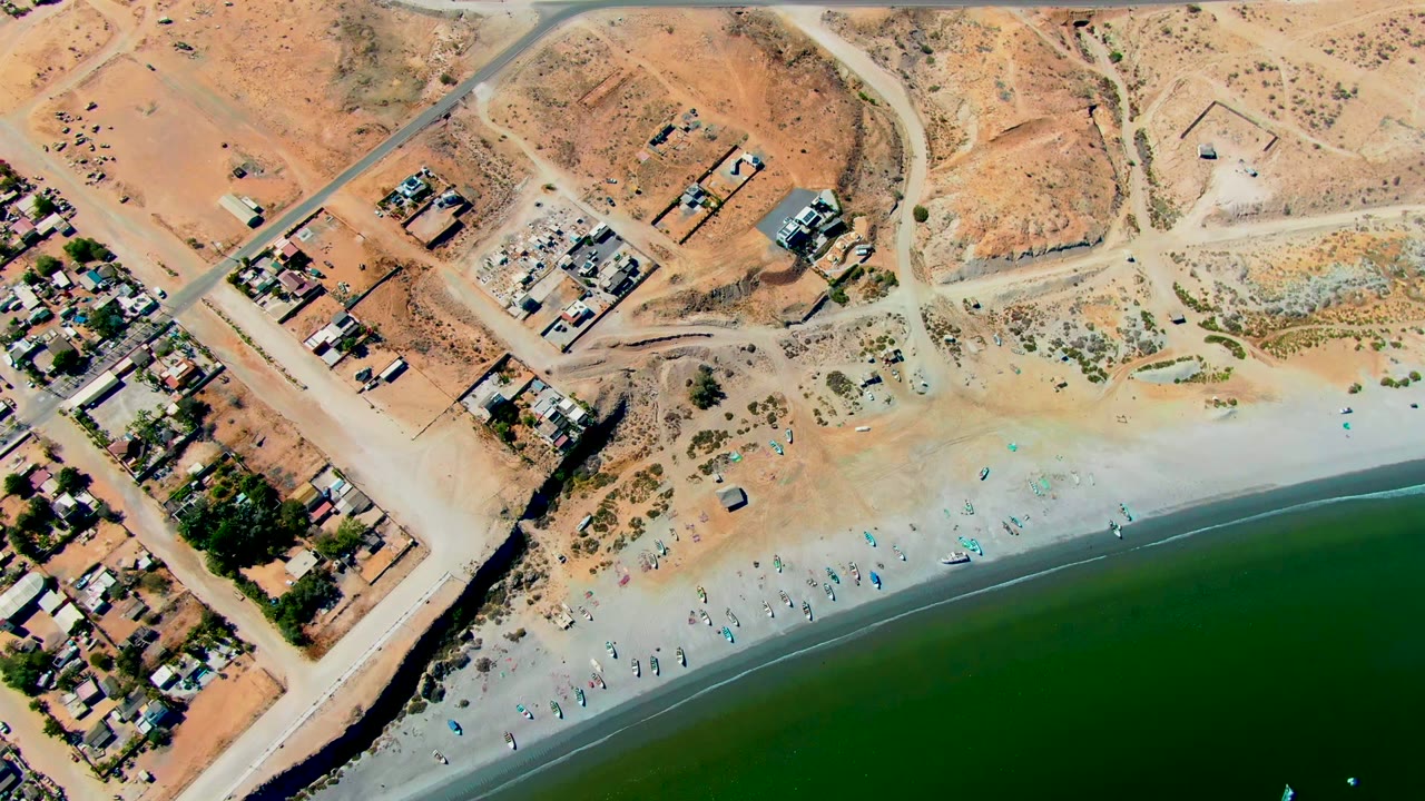 Aerial frame over San Juanico, Baja California Sur showing a paved road above dry lots, small buildings at left, a sandy beach with tiny people, and green water along the lower edge.