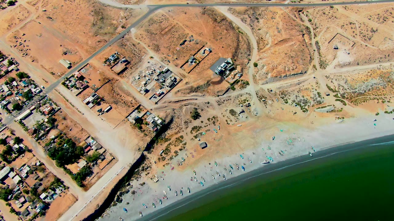 Drone view of sandy beach beside dry terrain and a road with scattered buildings.