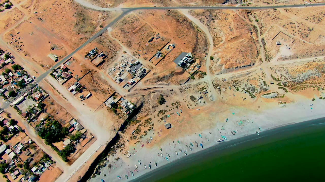 Aerial frame over San Juanico, Baja California Sur showing clustered small buildings on the left, a road crossing the upper frame, pale sand with scattered beachgoers, and dark green ocean water below.