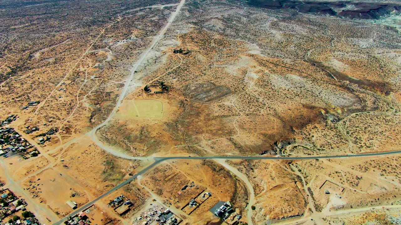 High aerial view over San Juanico, Baja California Sur, showing dry desert roads, scattered buildings, a pale beach, green ocean water, and small visible beachgoers near the shore.