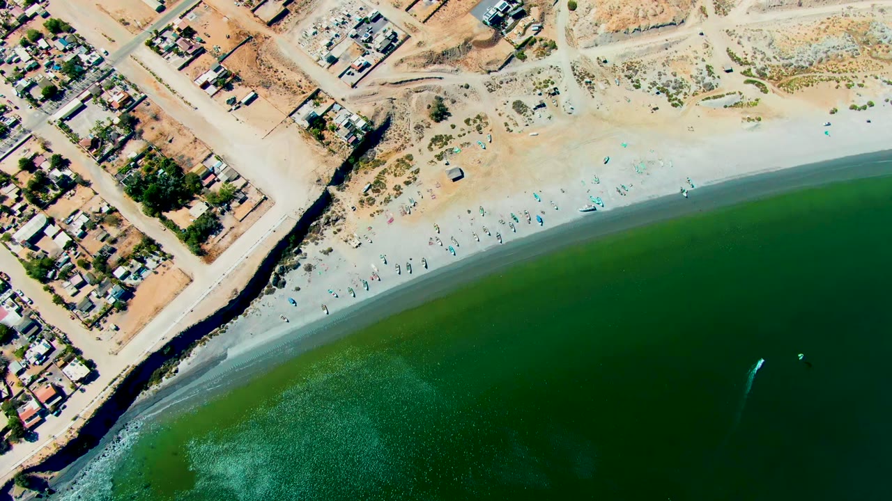 High overhead San Juanico frame showing more of the populated shoreline, with clustered buildings, a broad sandy beach, small people near the waterline, and green ocean below.