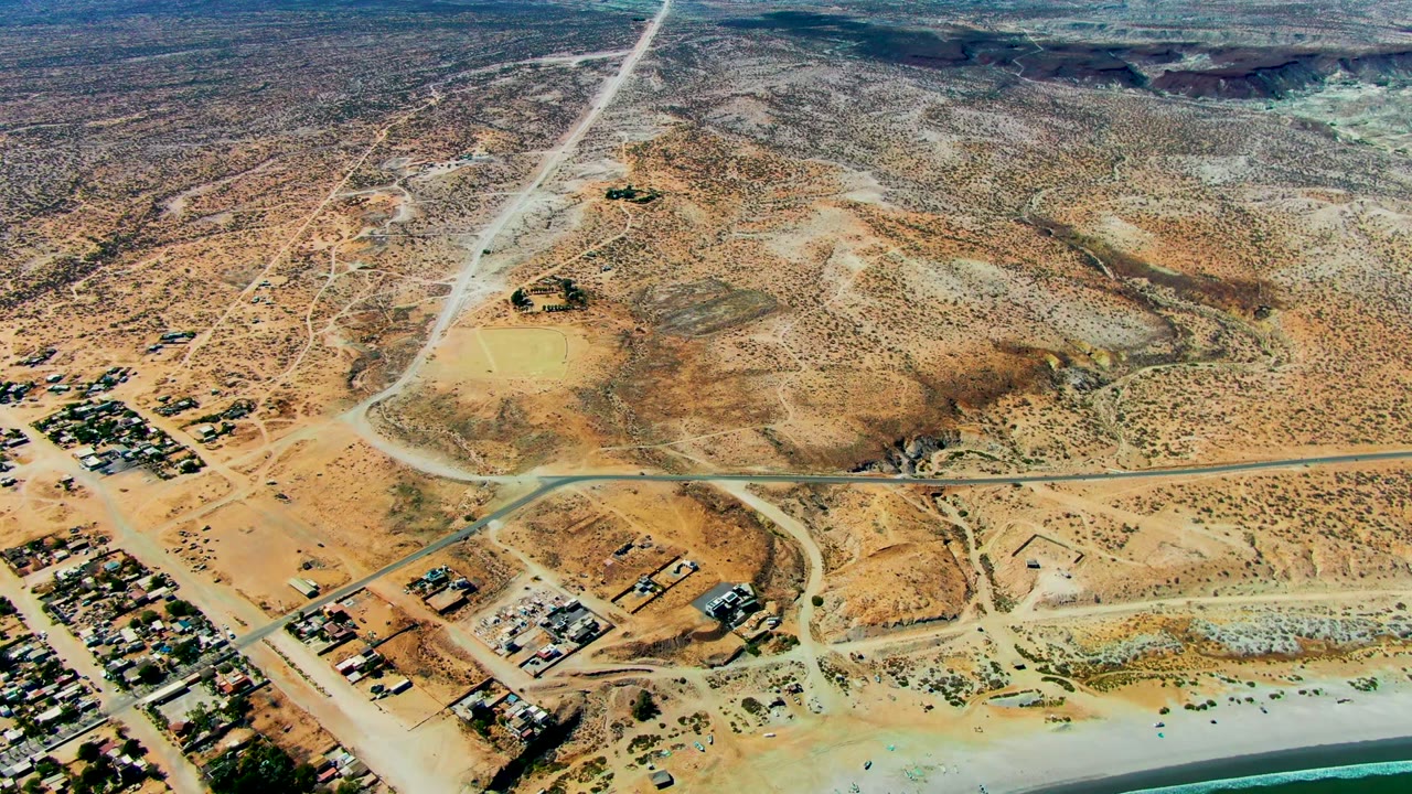Opening aerial frame over San Juanico, Baja California Sur, showing desert scrub, intersecting roads, small coastal building clusters, pale beach, turquoise water, and no visible people.