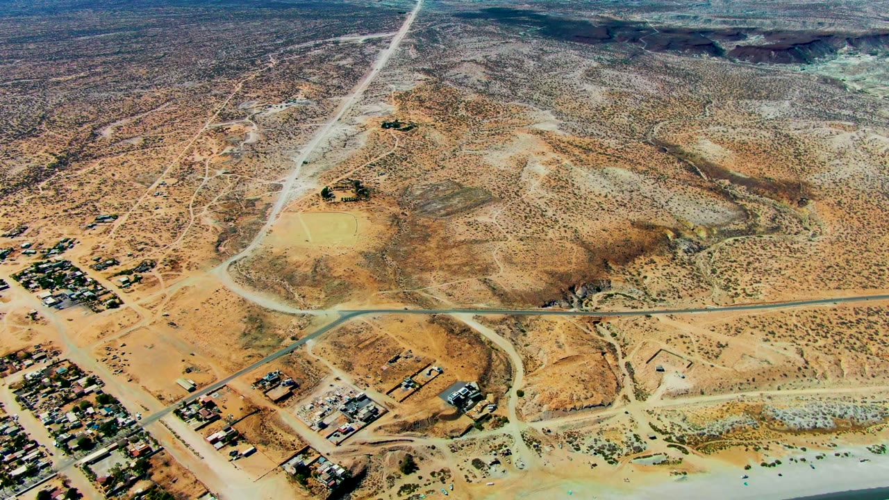 At 0:02, the frame shows San Juanico's sandy coastal edge, clustered low structures, diagonal roads crossing inland desert, a narrow surf line, and no visible people.