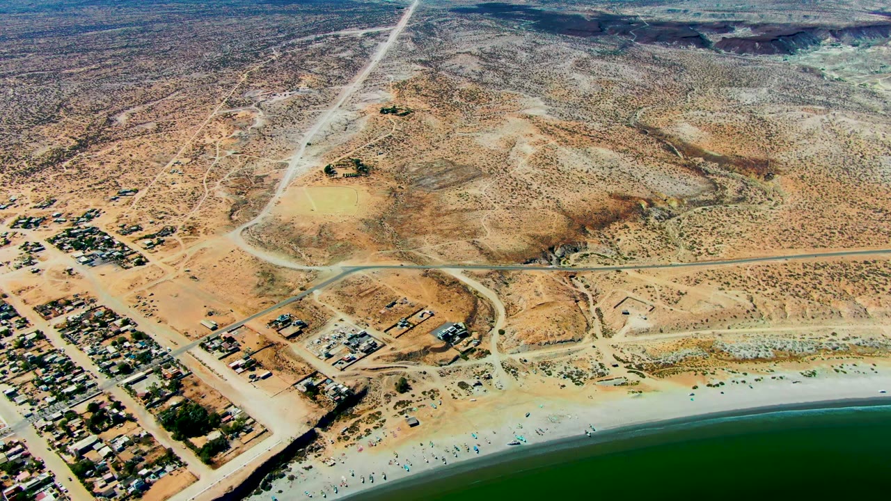 Aerial frame over San Juanico, Baja California Sur, showing green-blue ocean, pale beach with tiny visible people, nearby settlement blocks, dusty roads, and dry scrubland beyond.