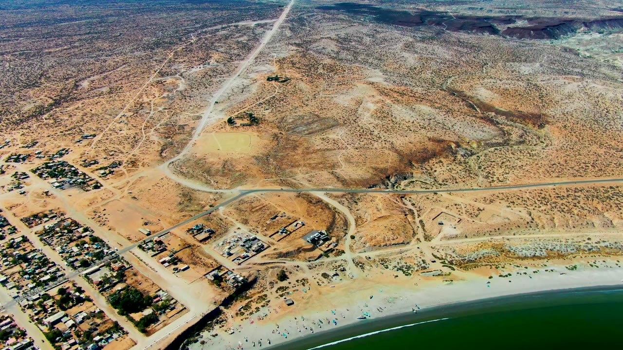 San Juanico aerial frame showing the ocean band at the bottom, a light sand beach with reduced visible activity, compact buildings near the coast, intersecting dirt roads, and broad desert terrain.
